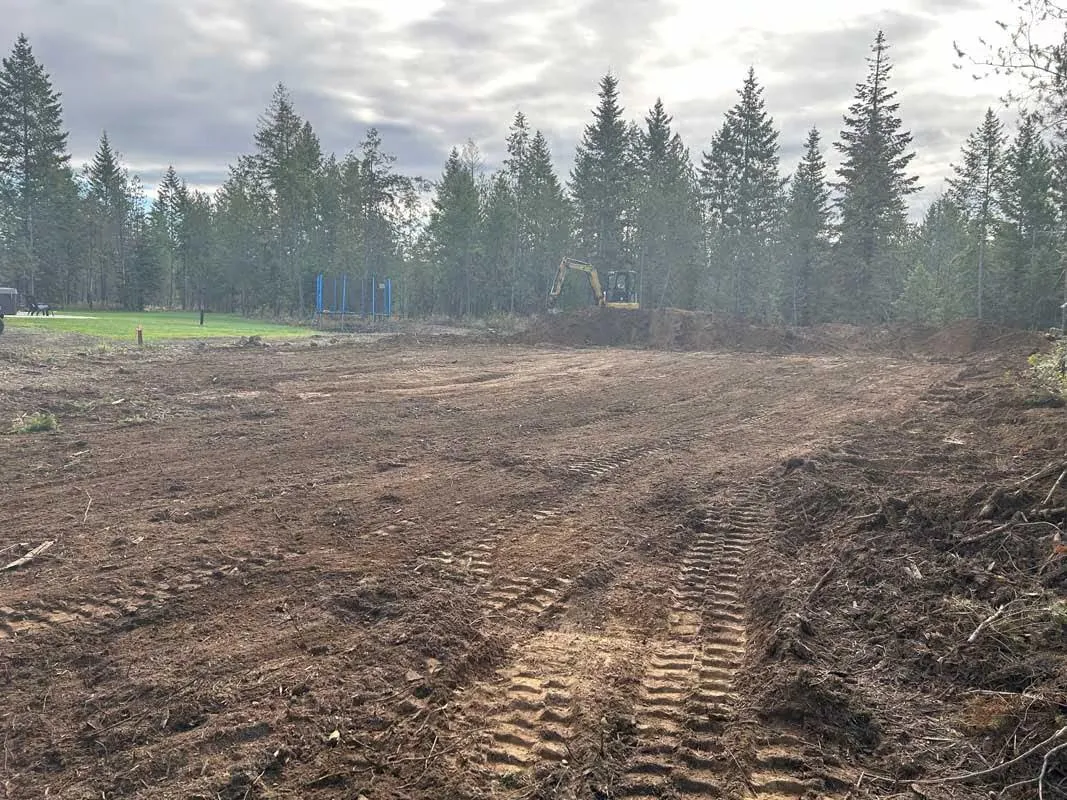 Cleared dirt field with track marks and an excavator working at the tree line