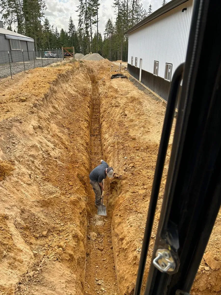 Worker shoveling in a deep utility trench dug beside a metal building, seen from an excavator cab